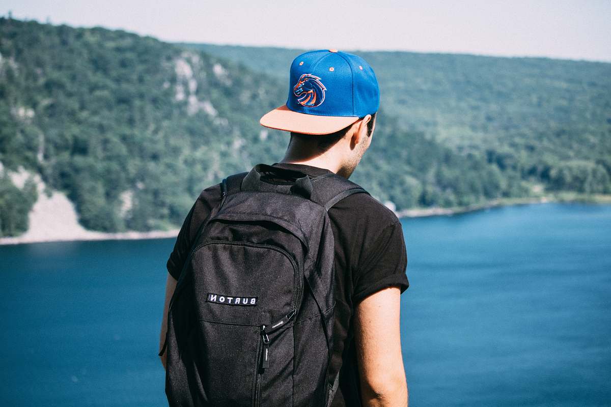 person man wearing black t-shirt, black backpack, and blue fitted cap facing body of water photo human