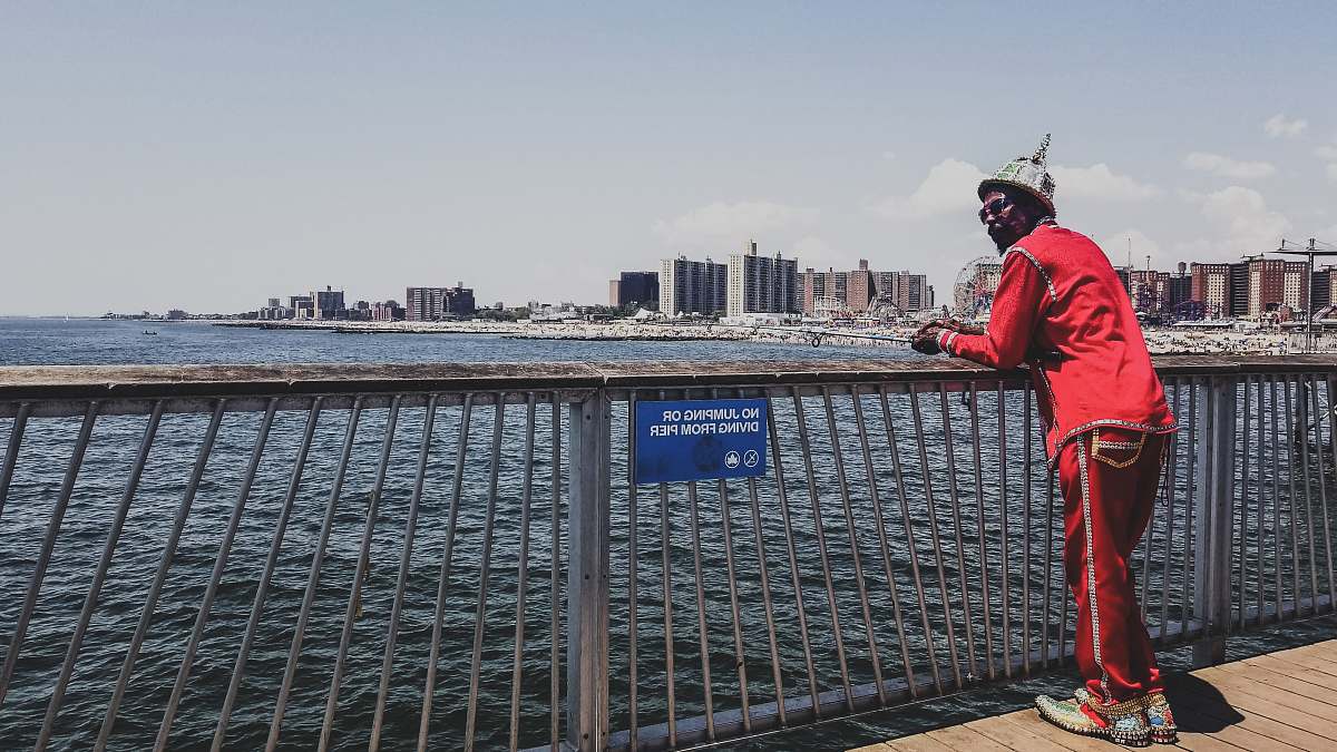 human man in red shirt standing and leaning on metal railings near sea during daytime person