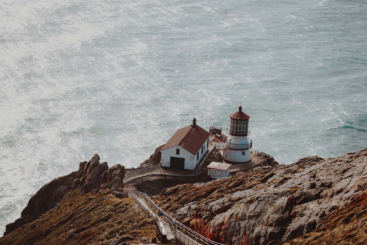 ocean aerial photography of white and brown concrete house beside lighthouse on gray mountain cliff near body of water at daytime outdoors