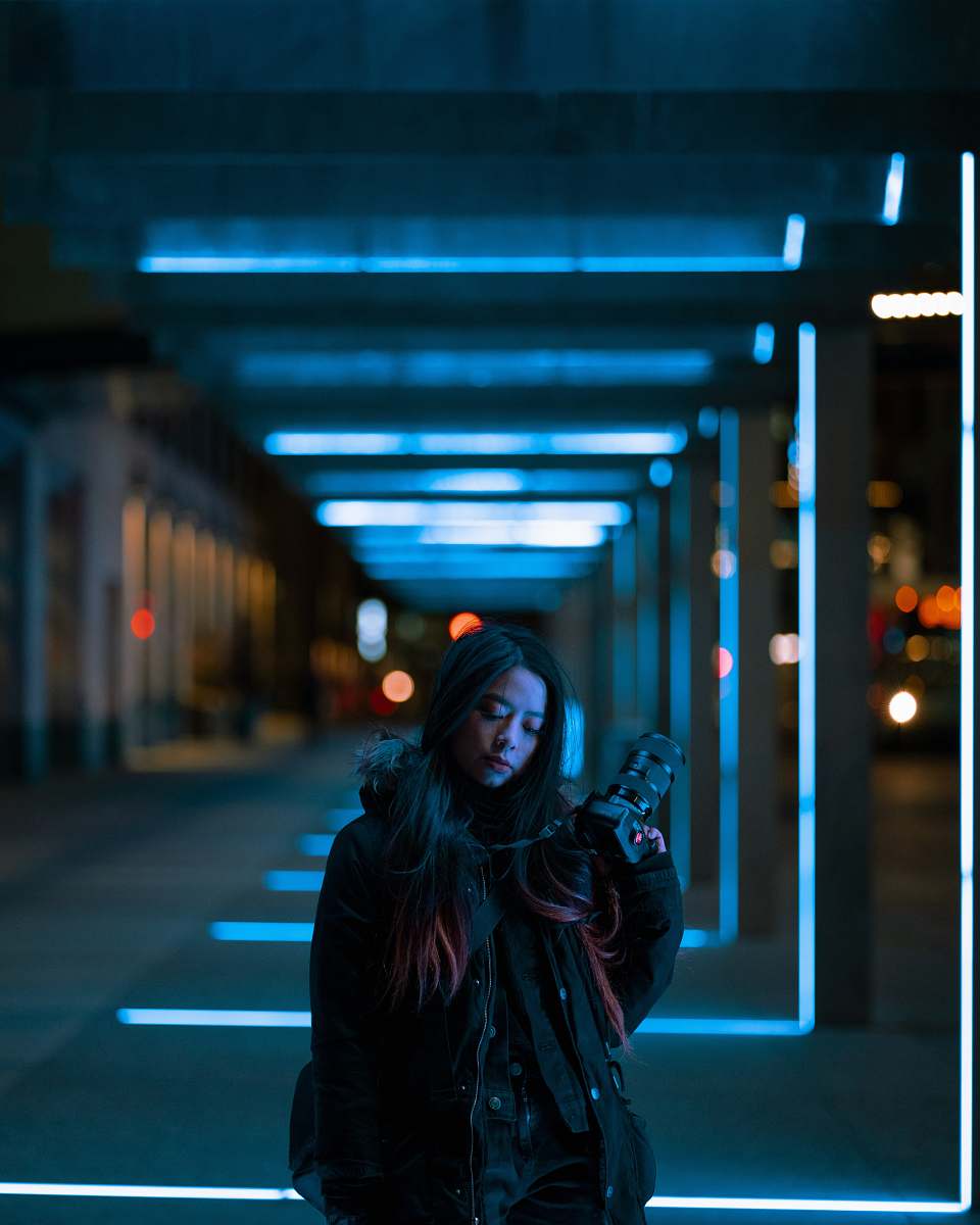 lighting woman in black leather jacket standing on sidewalk during night time apparel