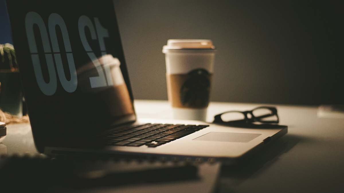 computer grey laptop computer displaying 12:00 on table near coffee cup and eyeglasses pc