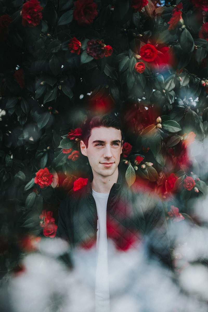 rose man in black zip-up jacket standing in frond of green plants and red flowers blossom
