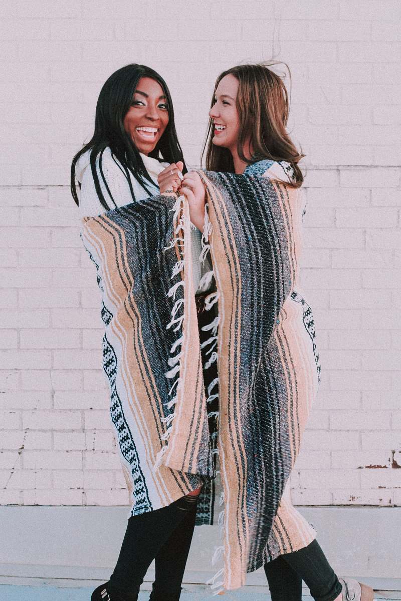 Capture the essence of friendship and warmth in this vibrant scene featuring a man and a woman happily covering themselves with a cozy blanket, while two women stroll down the street adorned in colorful ponchos. This image beautifully conveys the joy of companionship and the comfort of shared experiences, making it a perfect fit for lifestyle blogs, travel articles, or any content celebrating friendship and outdoor adventures.