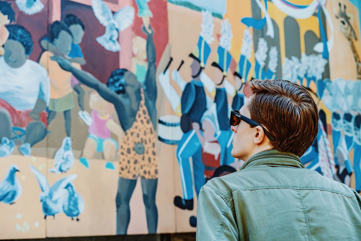 Capture the essence of urban culture with this striking image of a stylish man in a green collared top and sunglasses, standing confidently in front of a vivid graffiti mural. This scene not only showcases contemporary street fashion but also reflects the vibrant spirit of city life. Ideal for use in articles about fashion, urban culture, or art, this image serves as a perfect visual for any project that aims to convey energy and creativity.