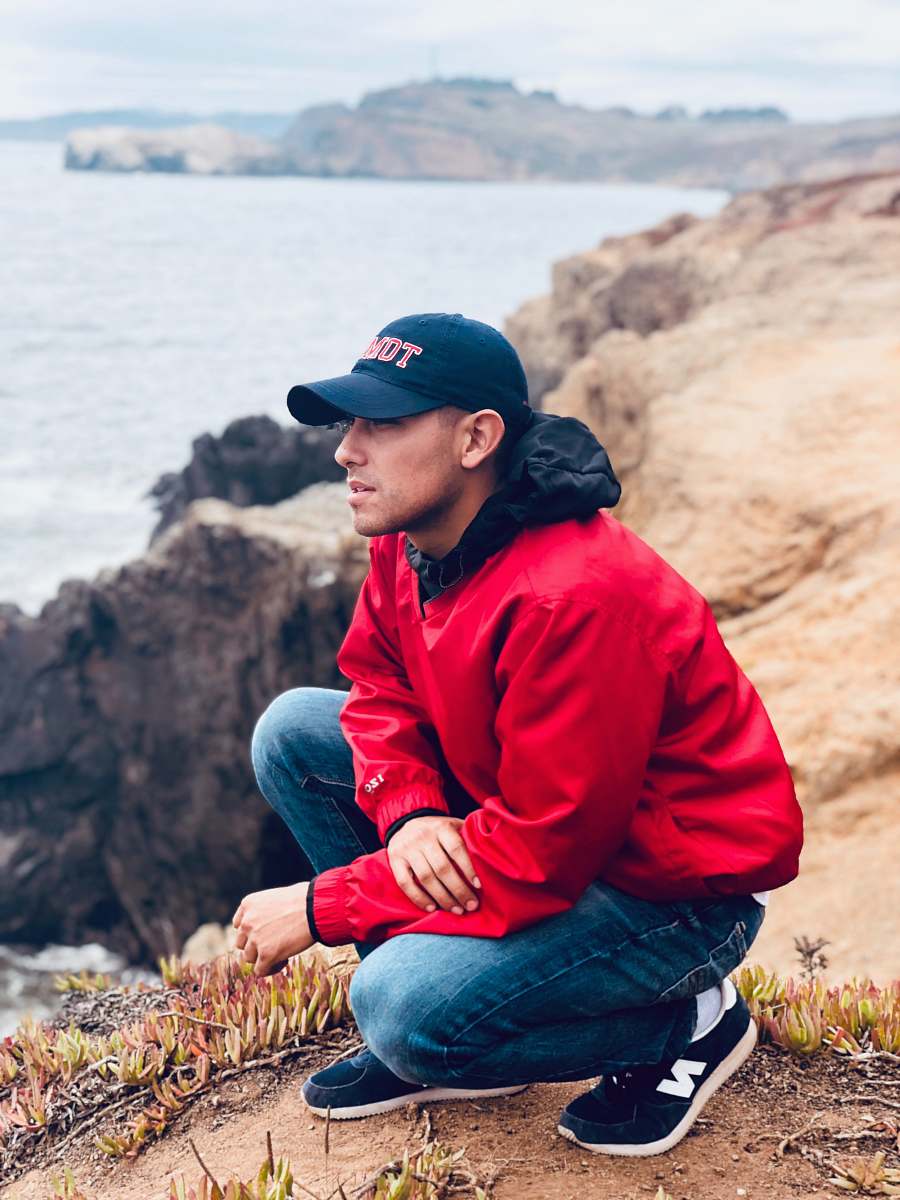 person man wearing cap and jacket squatting on top of the cliff looking the overview people