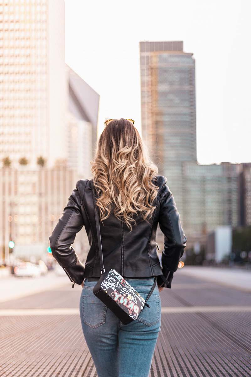 person woman wearing black leather jacket and blue denim bottoms standing on gray concrete surface near buildings during daytime people