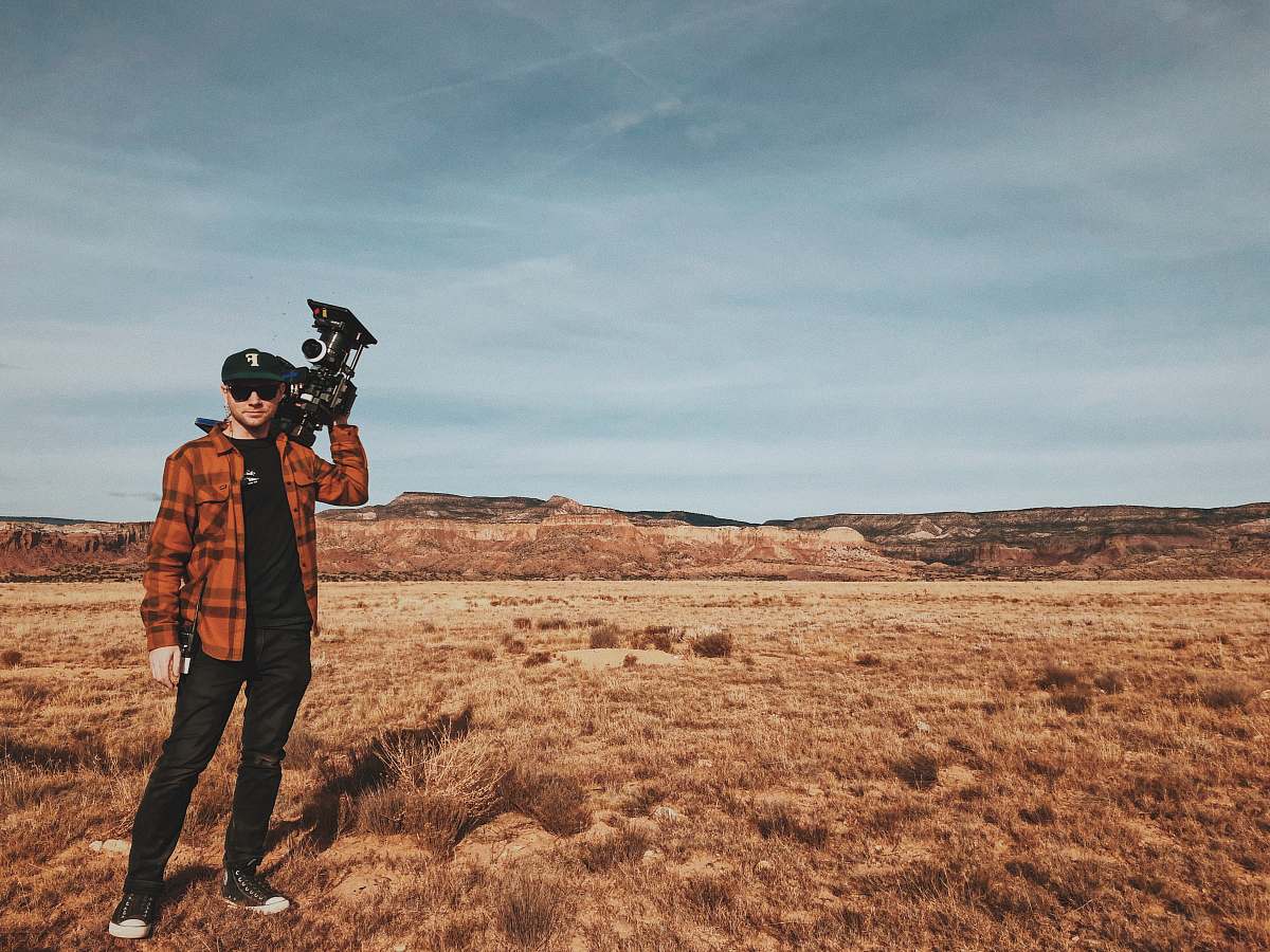 human man standing on brown field carrying black recording camera during daytime people