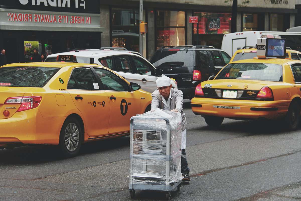 cab man pushing gray bus cart on road near two yellow taxi cabs car