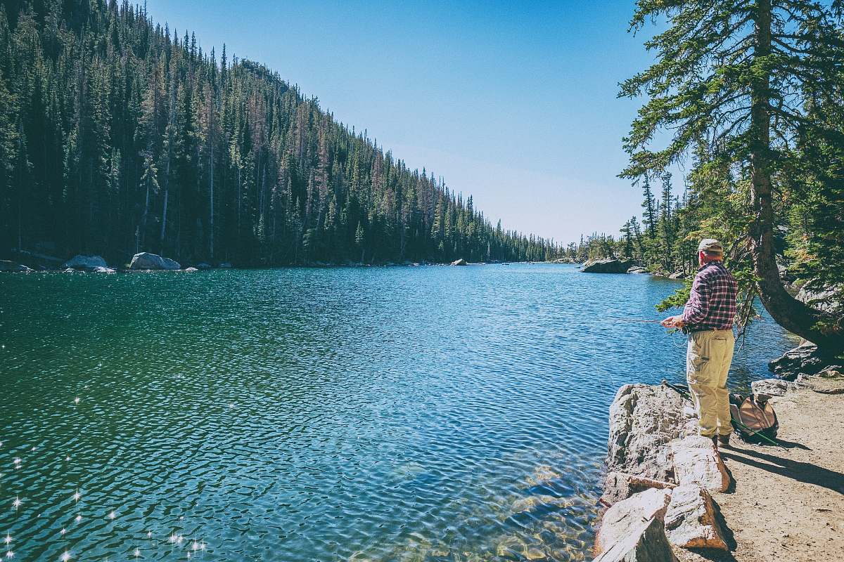 lake man standing on rock on front of lake surrounded with trees at daytime fishing