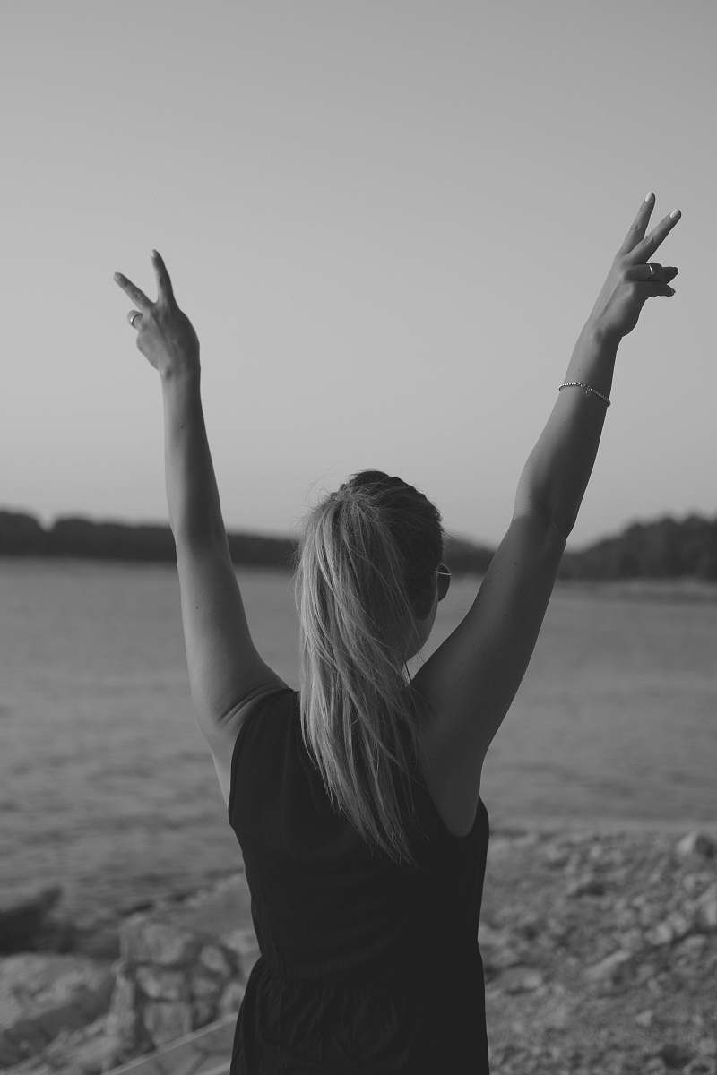 child grayscale photo of woman wearing black tank top walking on seashore while doing peace hand sign teen