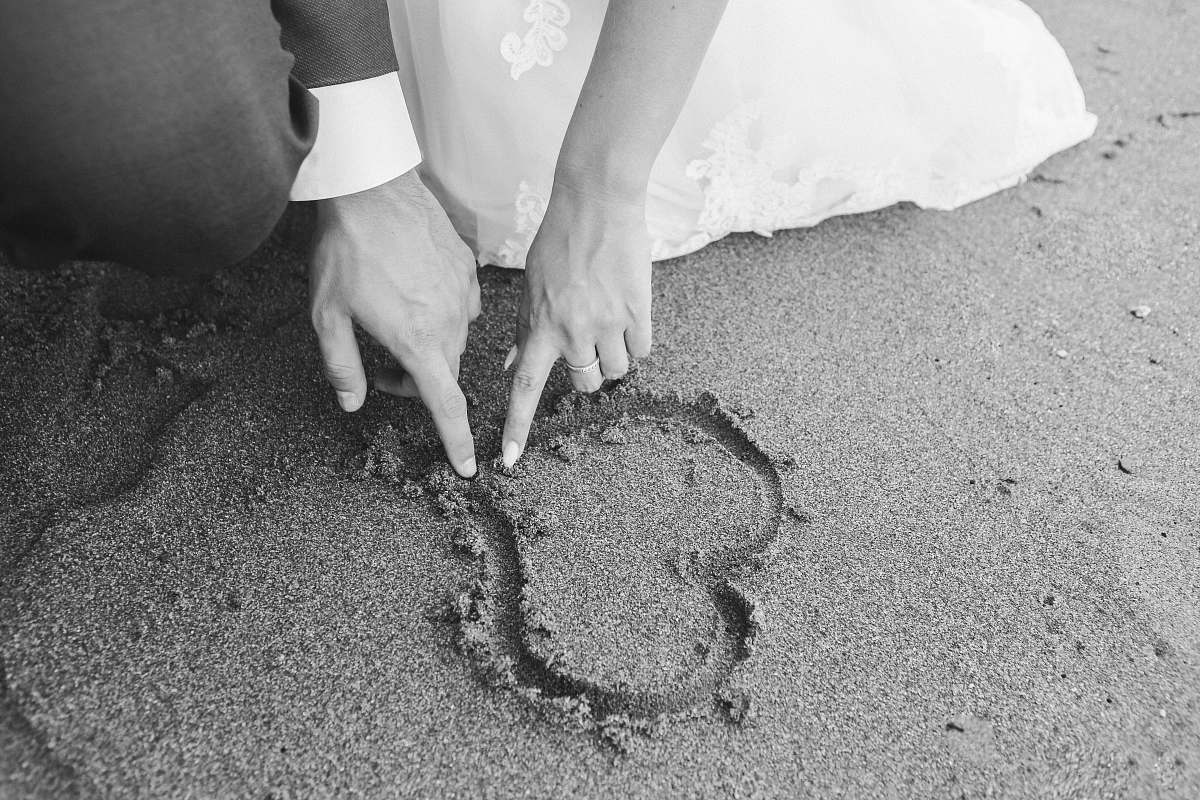 grey grayscale photography of man and woman draw heart on sand wedding