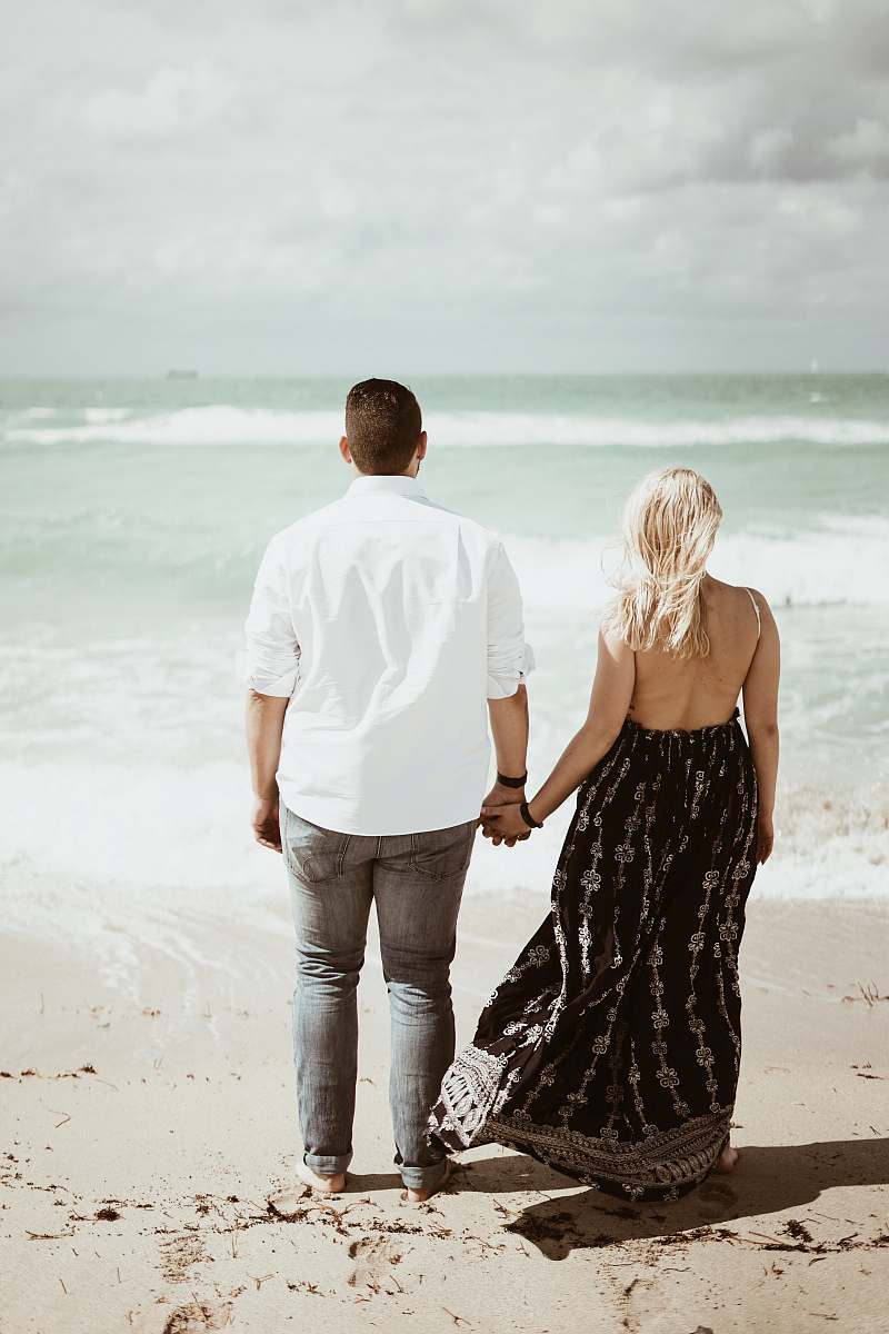 person man and woman holding each other's hands and walking by the seashore during daytime clothing