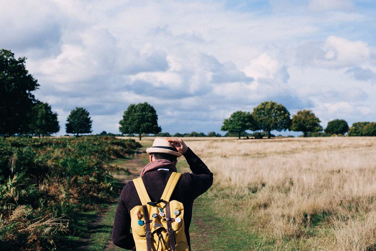 person man holding fedora cap while walking on green and beige grass field under white clouds people