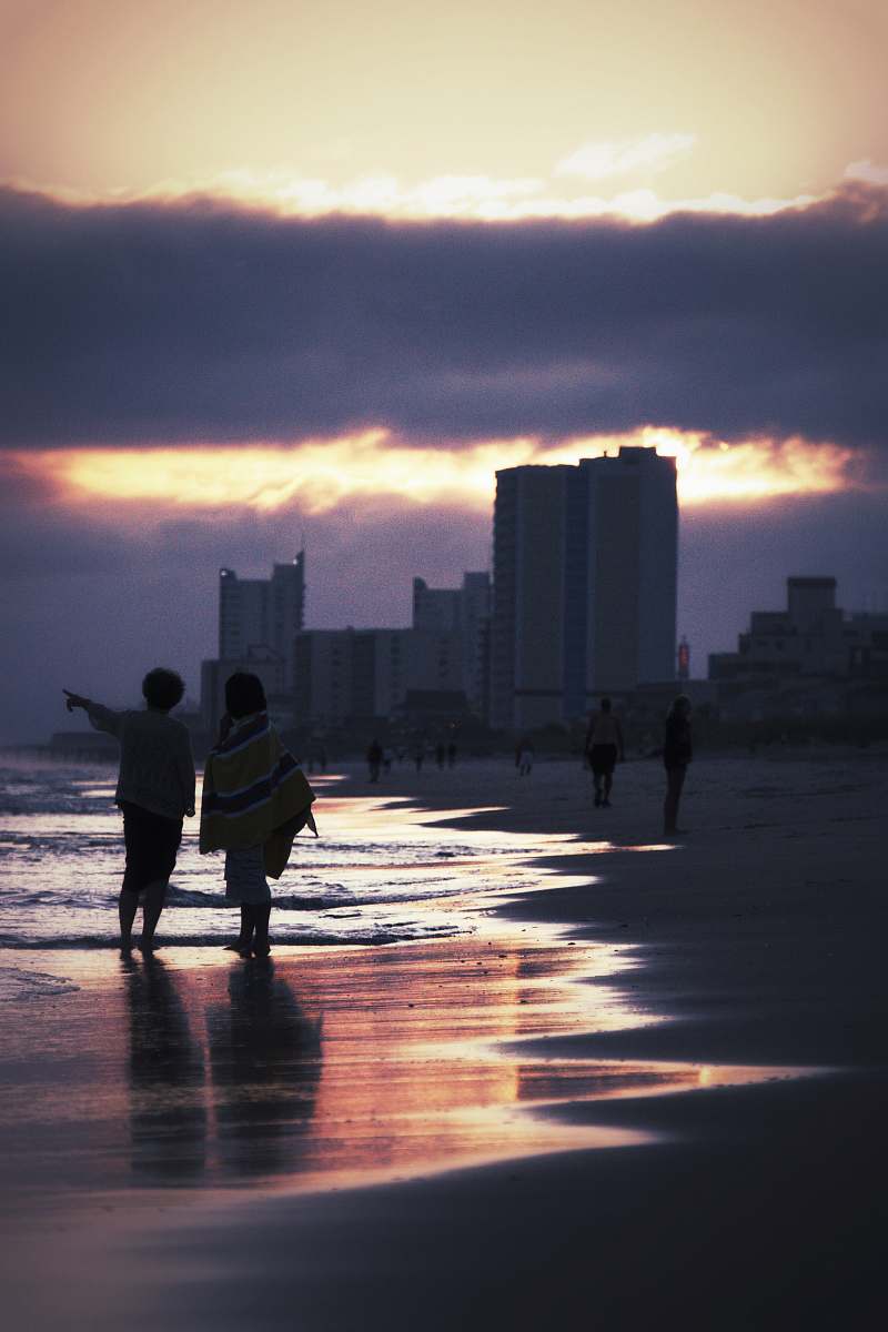 person two people walking on seashore near buildings during golden hour silhouette