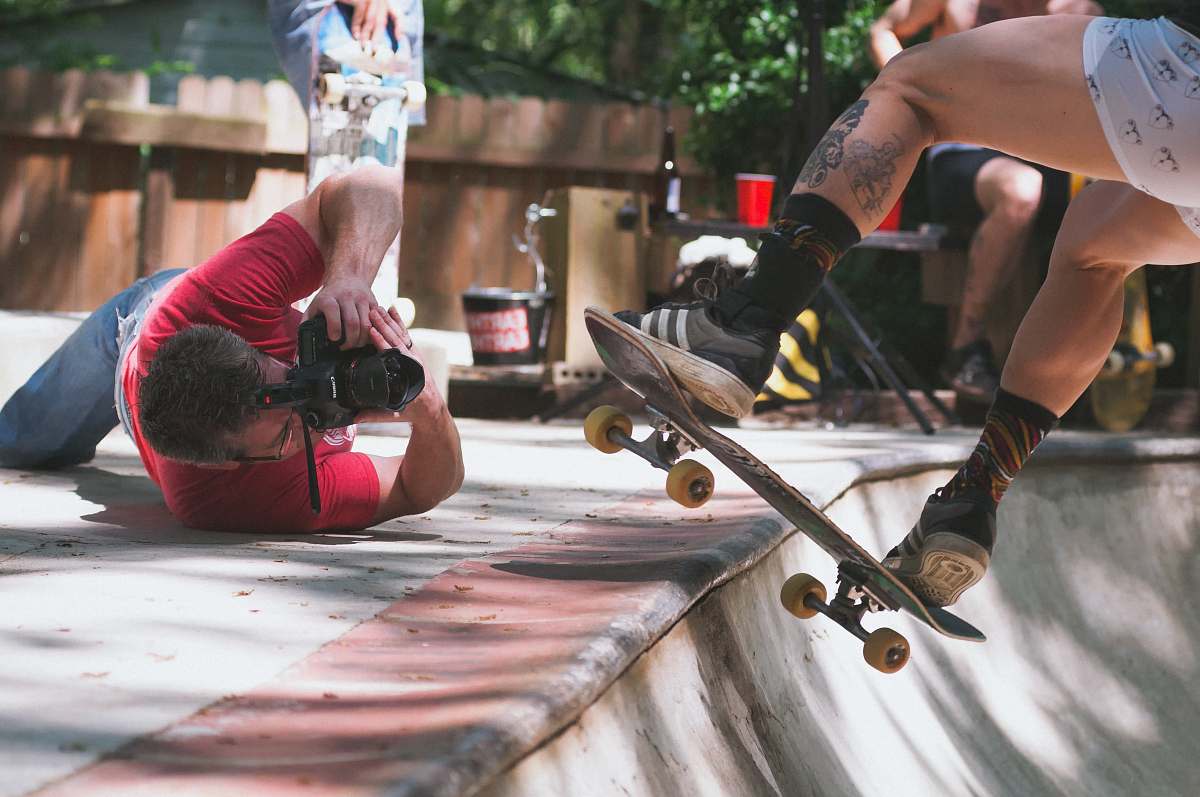 human man lying on floor while taking picture of person doing skate trick on ramp photographer