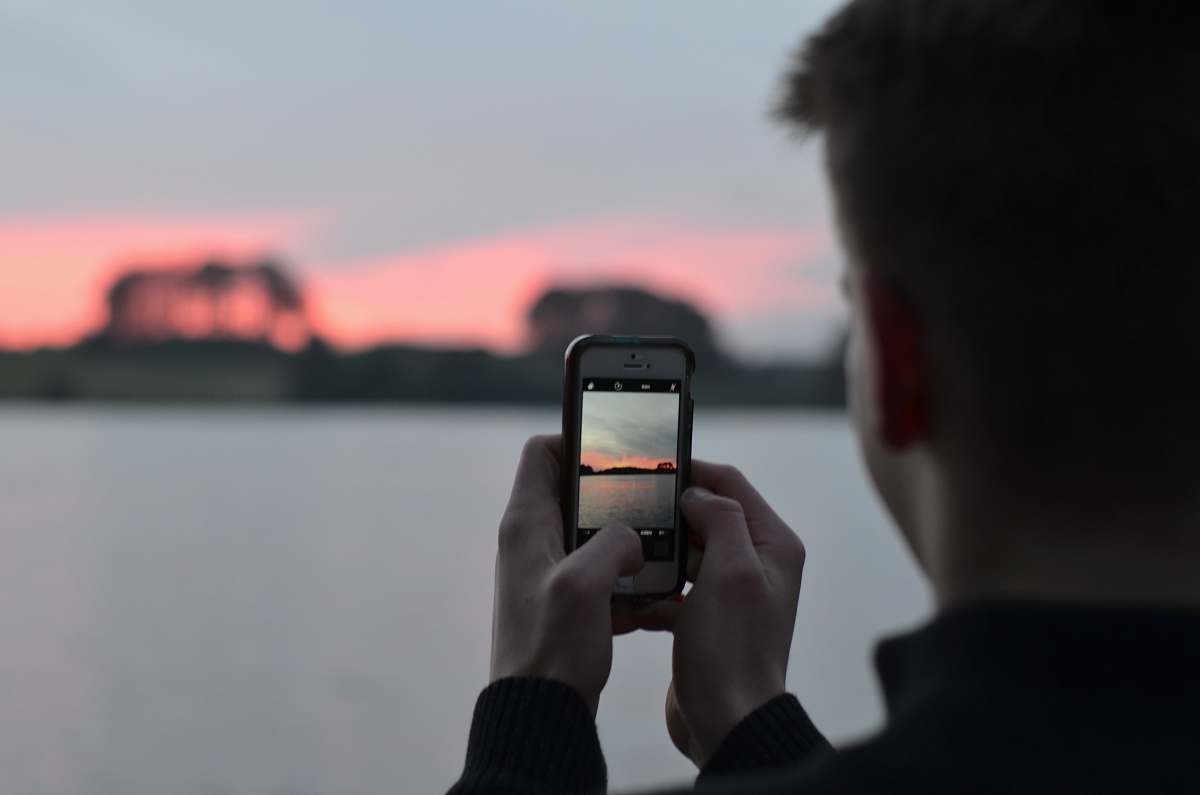 human selective focus photography of person using smartphone camera taking photo of blue body of water people
