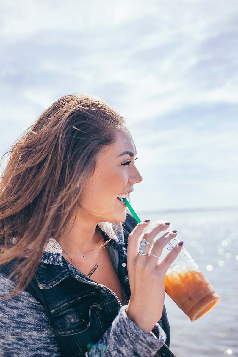 people woman wearing black and grey jacket holding glass cup while drinking near body of water during daytime smiling