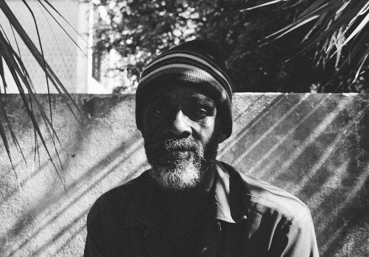 beard grayscale photo of man wearing beanie in front of concrete wall and between two linear-leafed plants portrait
