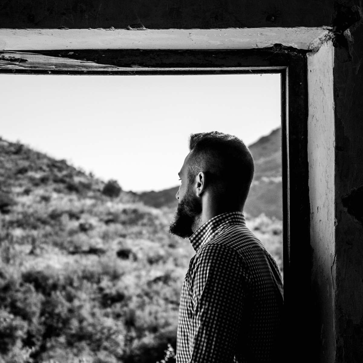 people grayscale photography of man in tattersall top looking outside of window person