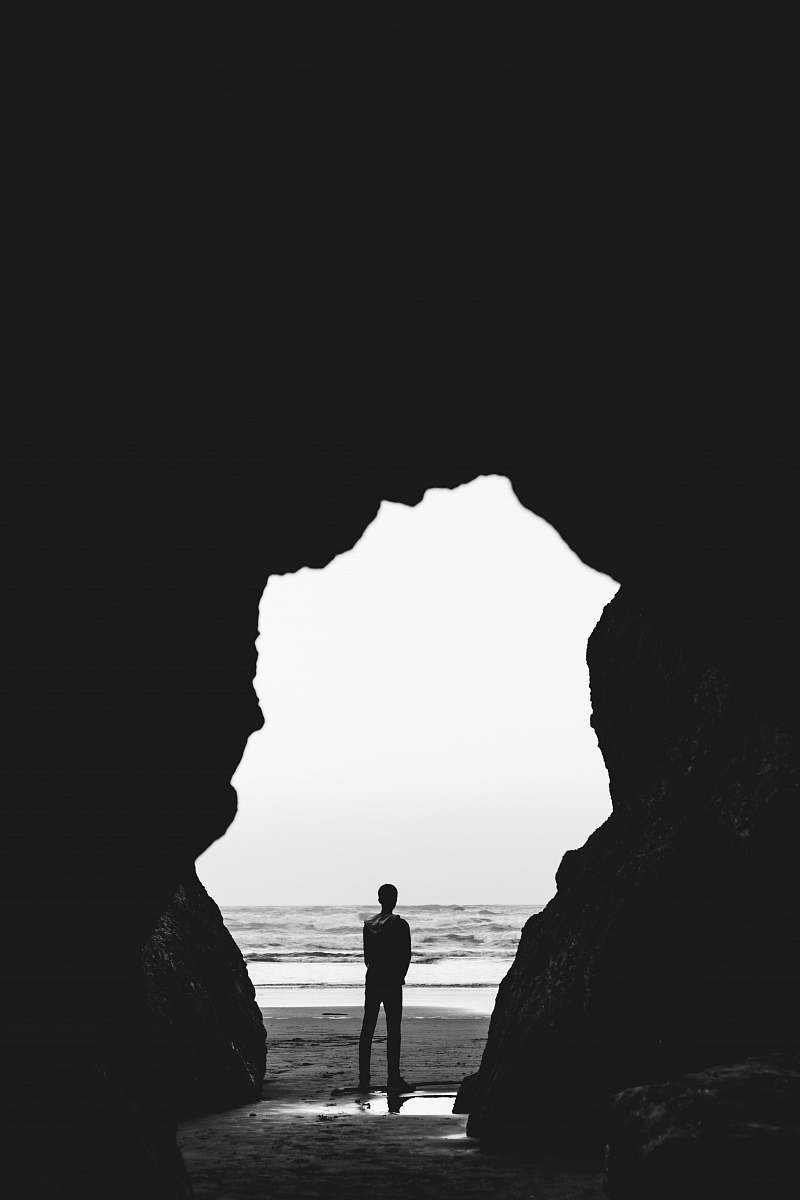 cave grayscale photography of man standing on seashore ocean