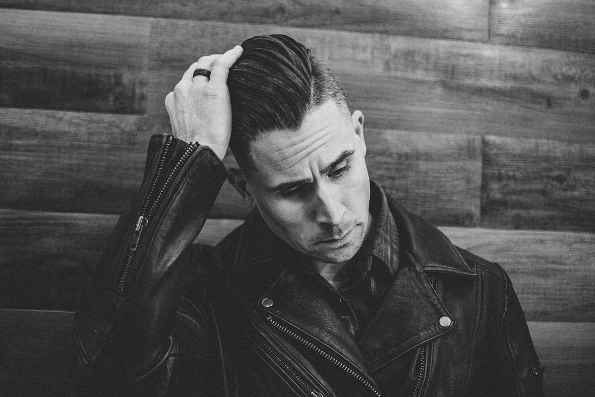 person grayscale photography of man wearing leather jacket leaning against wall while fixing hair people