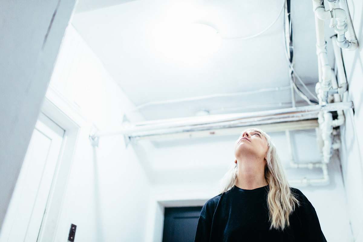 A striking image of a blonde woman dressed in a sleek black crew-neck shirt, standing in a softly lit room with her head tilted upwards. Her gaze is directed at the ceiling, evoking a sense of curiosity and contemplation. Perfect for use in articles related to introspection, personal growth, or lifestyle branding, this image captures a moment of thoughtful reflection that resonates with a broad audience.