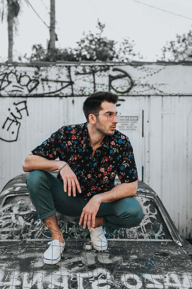 person man in black, blue, and red floral button-up collared shirt and gray pants sitting on car hood during daytime people