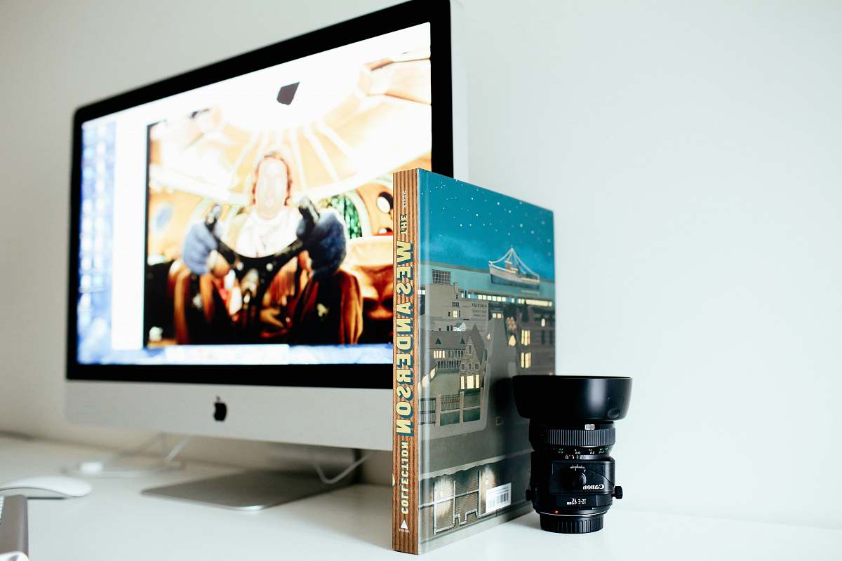 screen selective focus photography of black camera lens beside book and turned on iMac office