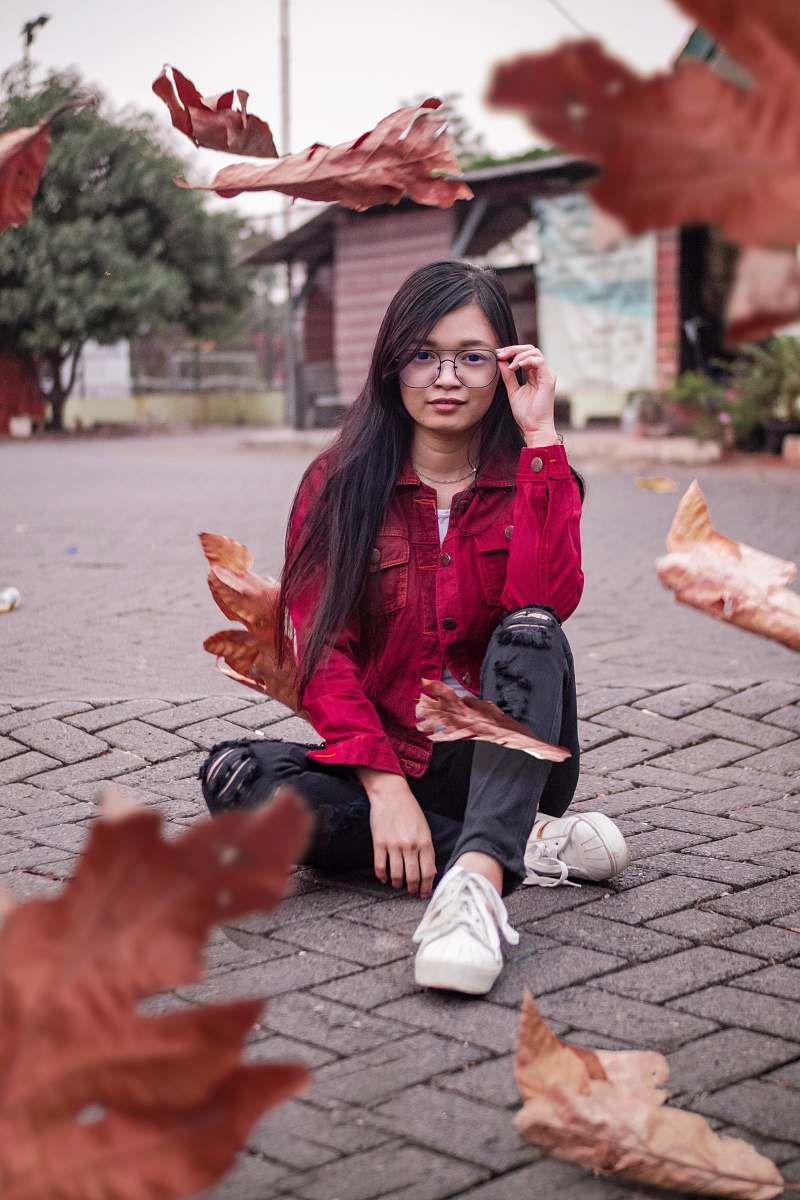 clothing woman wearing red denim jacket and black skinny jeans sitting on concrete pavement holding eyeglasses footwear