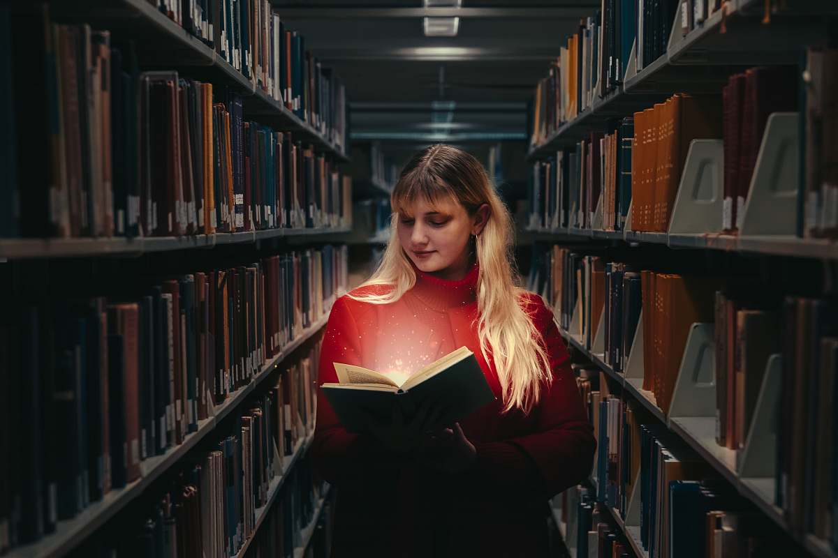 library woman in red turtleneck sweater holding book between library shelves indoors
