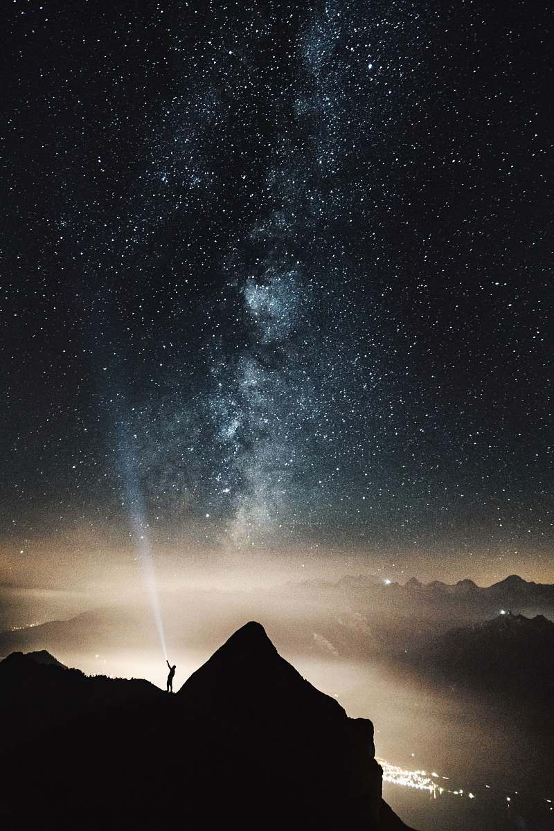 star silhouette of person on top of mountain pointing flashlight on sky filled with stars at night time milky way