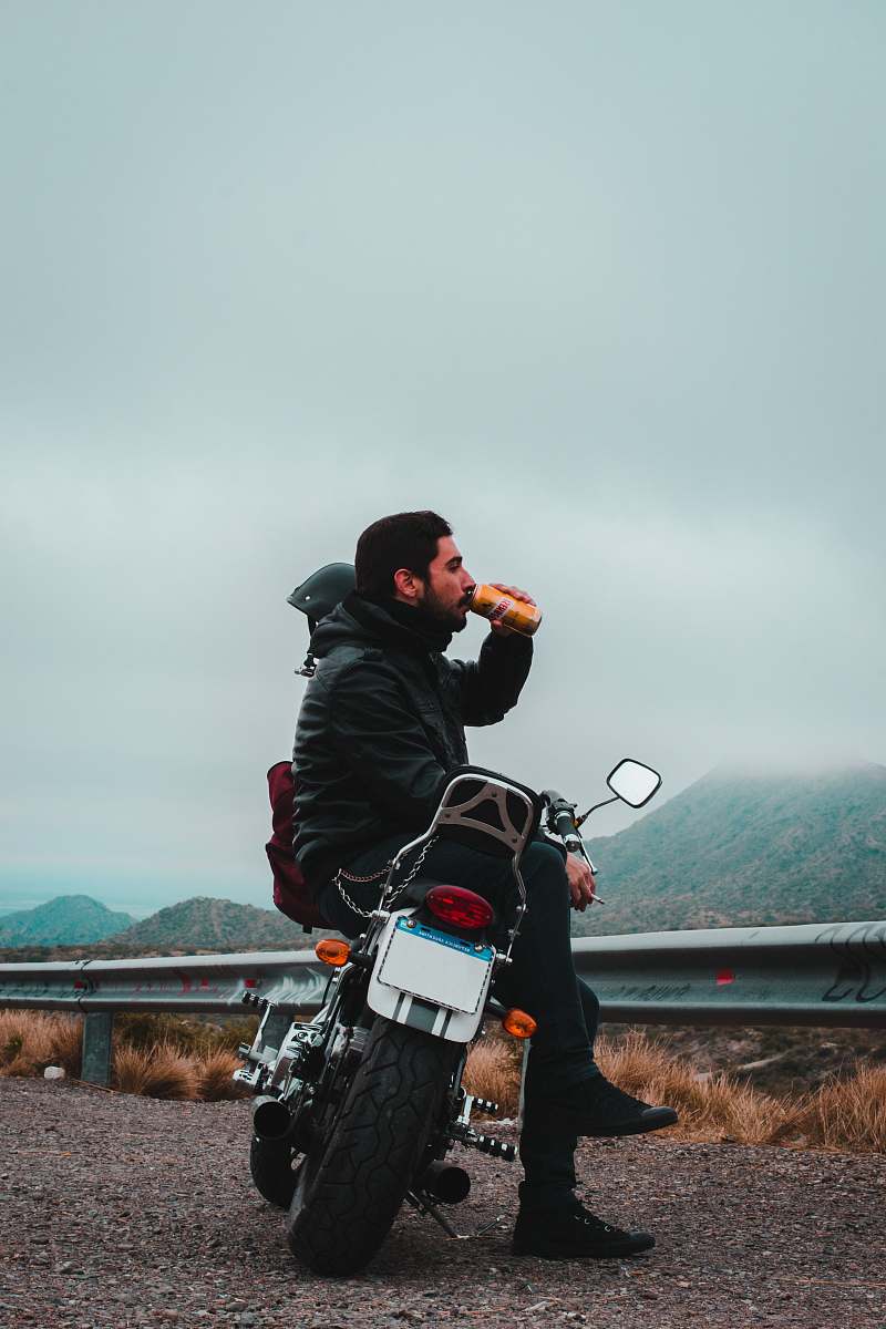 human man sitting on motorcycle while drinking beverage in can overlooking mountain under dark cloudy skies at daytime motorcycle