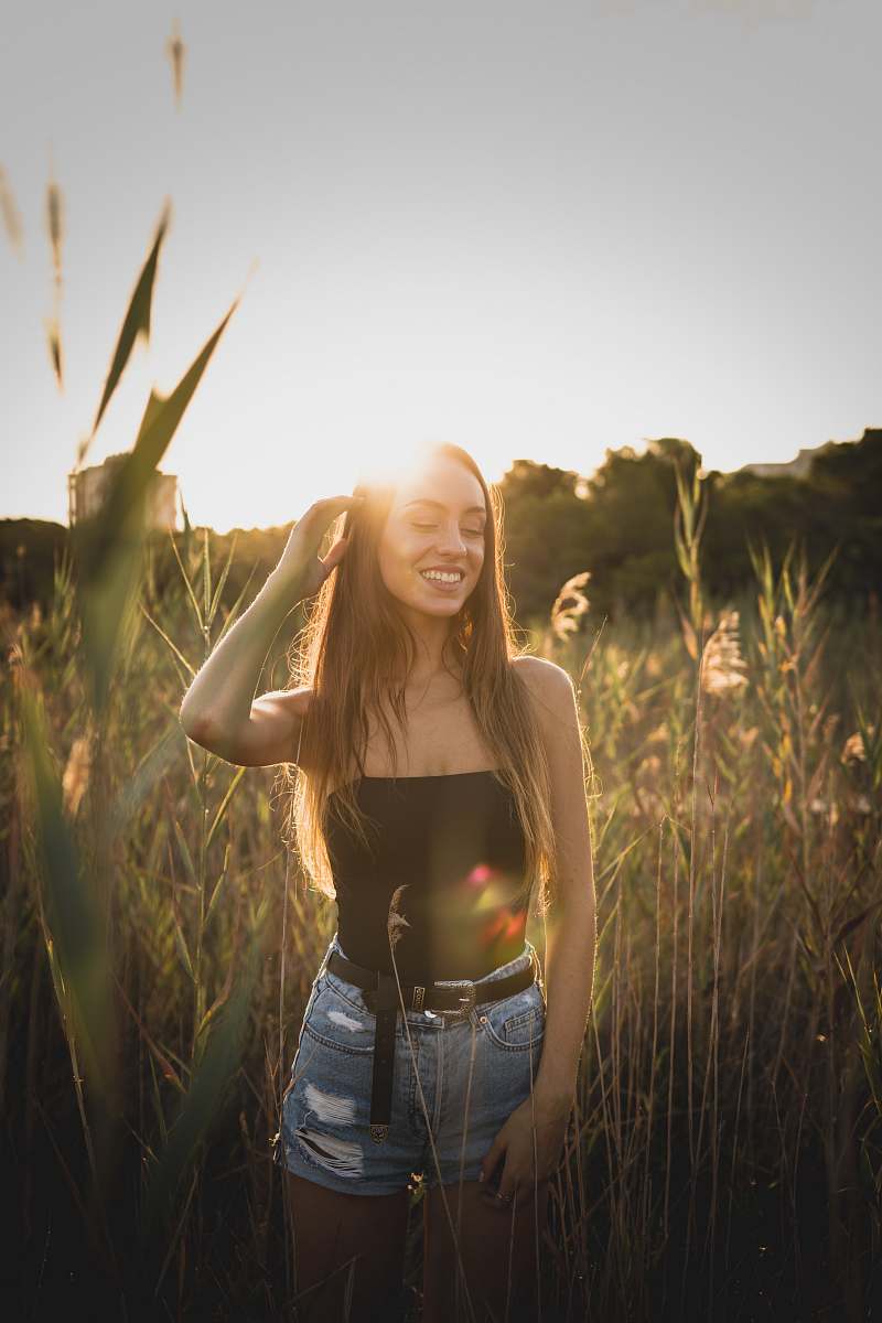 human smiling woman in black strapless top and blue distressed shorts standing near green grass during sunset people