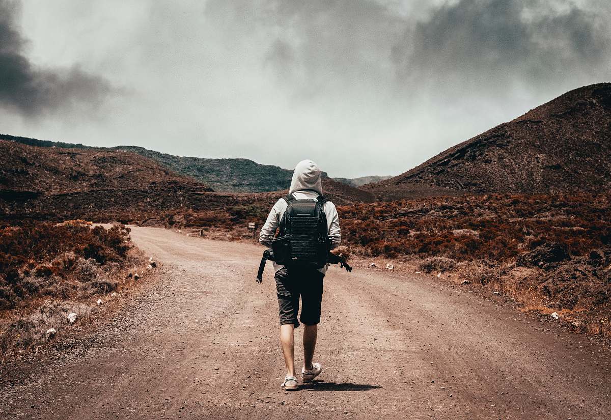gravel man in gray hoodie carrying backpack walking under the sun dirt road