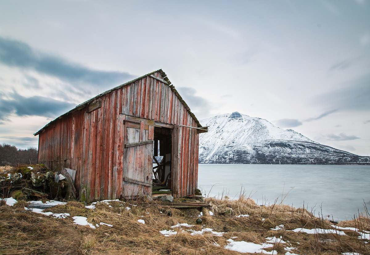 hut landscape photography of brown wooden shed near ocean water and snow-coated mountains during daytime rural