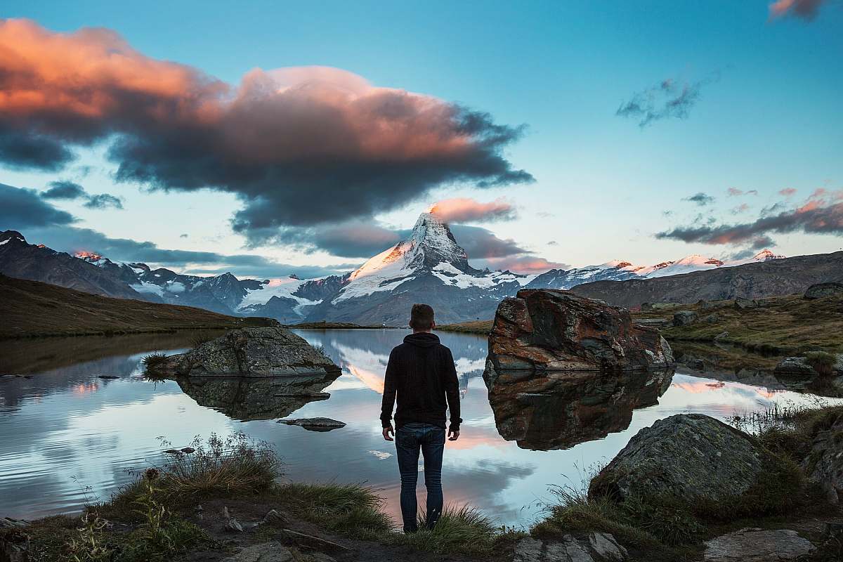 mountain man standing on rocky cliff facing body of water alps