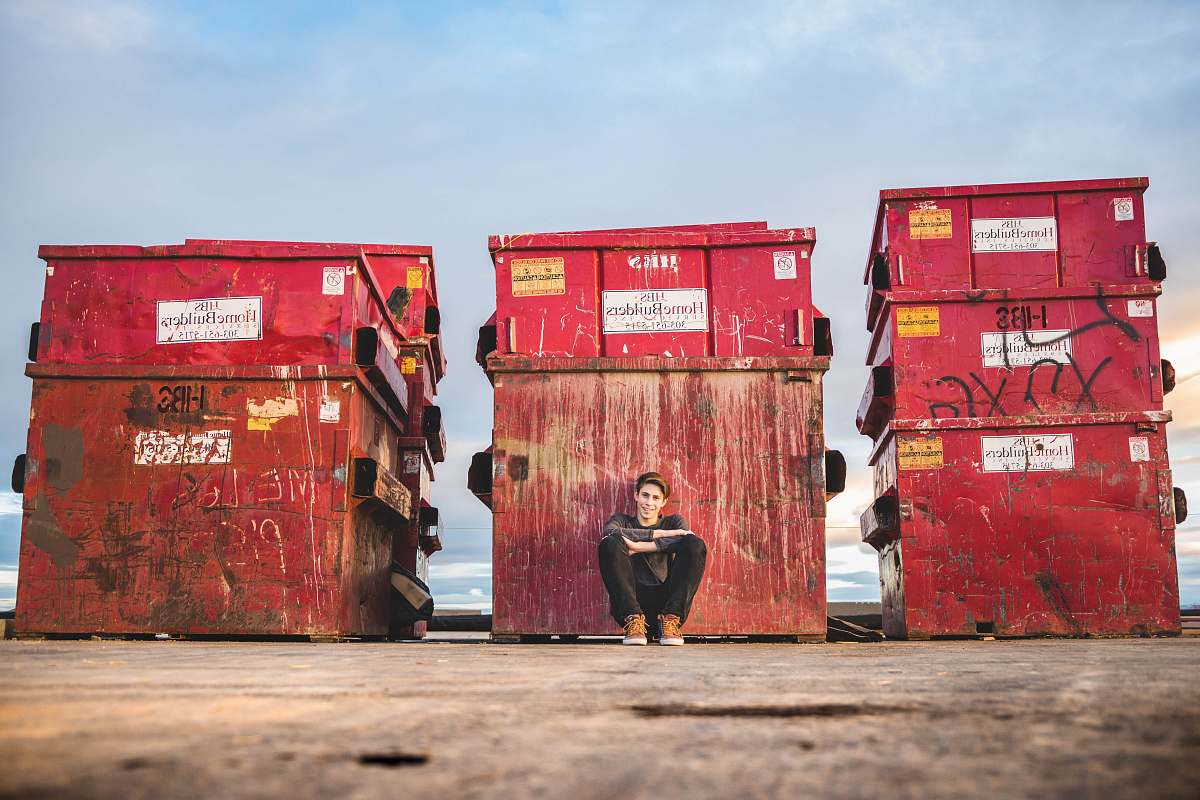 In this striking urban scene, a young boy leans casually while seated on a box amidst vibrant graffiti, embodying the spirit of city life. Next to him, a red trash bin adds a pop of color, creating a juxtaposition between art and the everyday environment. This image is perfect for projects related to urban culture, youth lifestyle, street art, and environmental themes, making it a versatile addition to your collection.