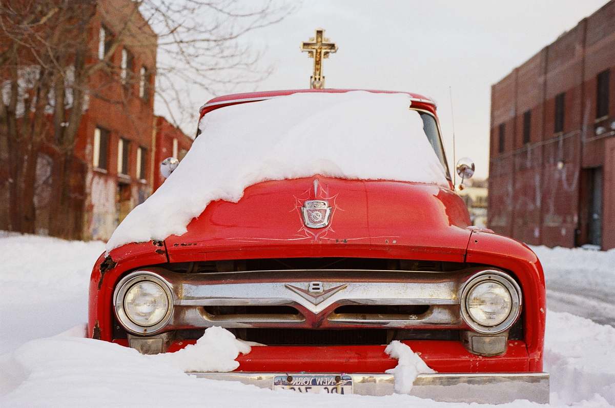 car red vehicle surrounded by snowy field beside brown concrete buildings during daytime vehicle