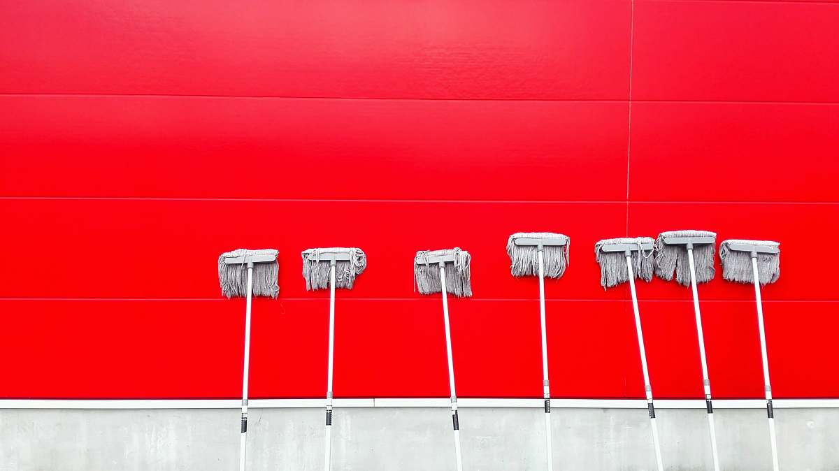 This vibrant image features a striking arrangement of seven white push mops neatly lined up against a vivid red wall. The sharp contrast between the clean white mops and the dynamic color of the wall creates an eye-catching visual ideal for cleaning service promotions, home improvement blogs, or product catalogs. Perfect for illustrating cleanliness, organization, and professional cleaning services.