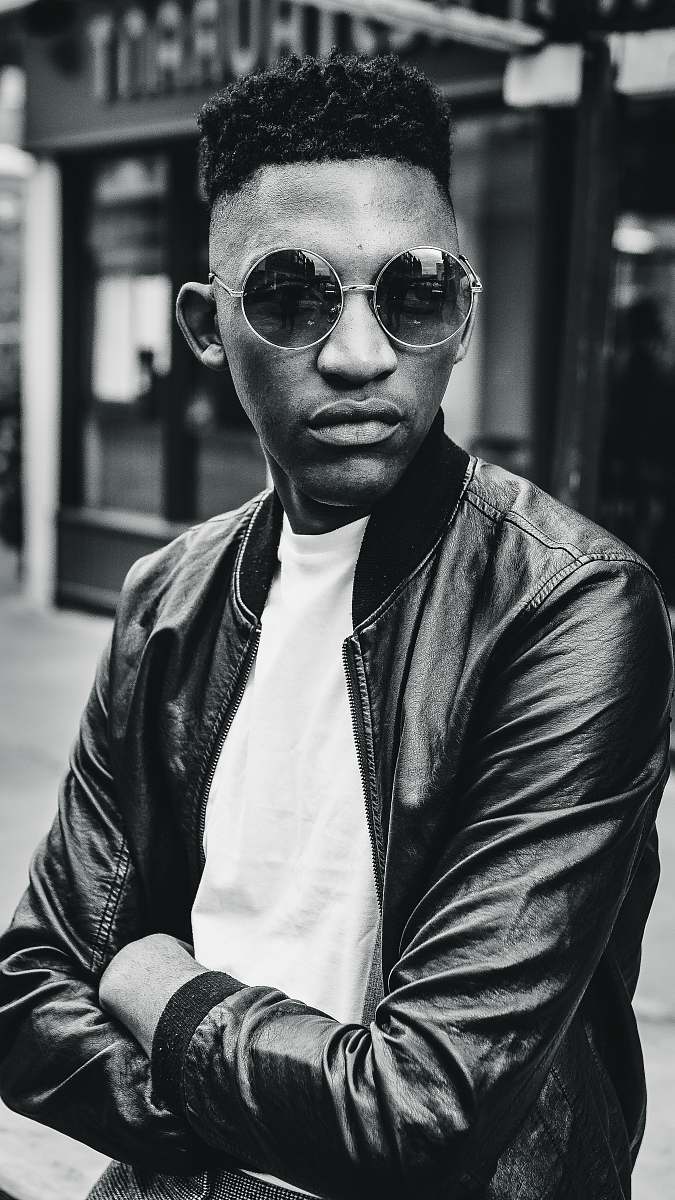 people grayscale photo of man wearing bomber jacket standing outdoor during daytime person