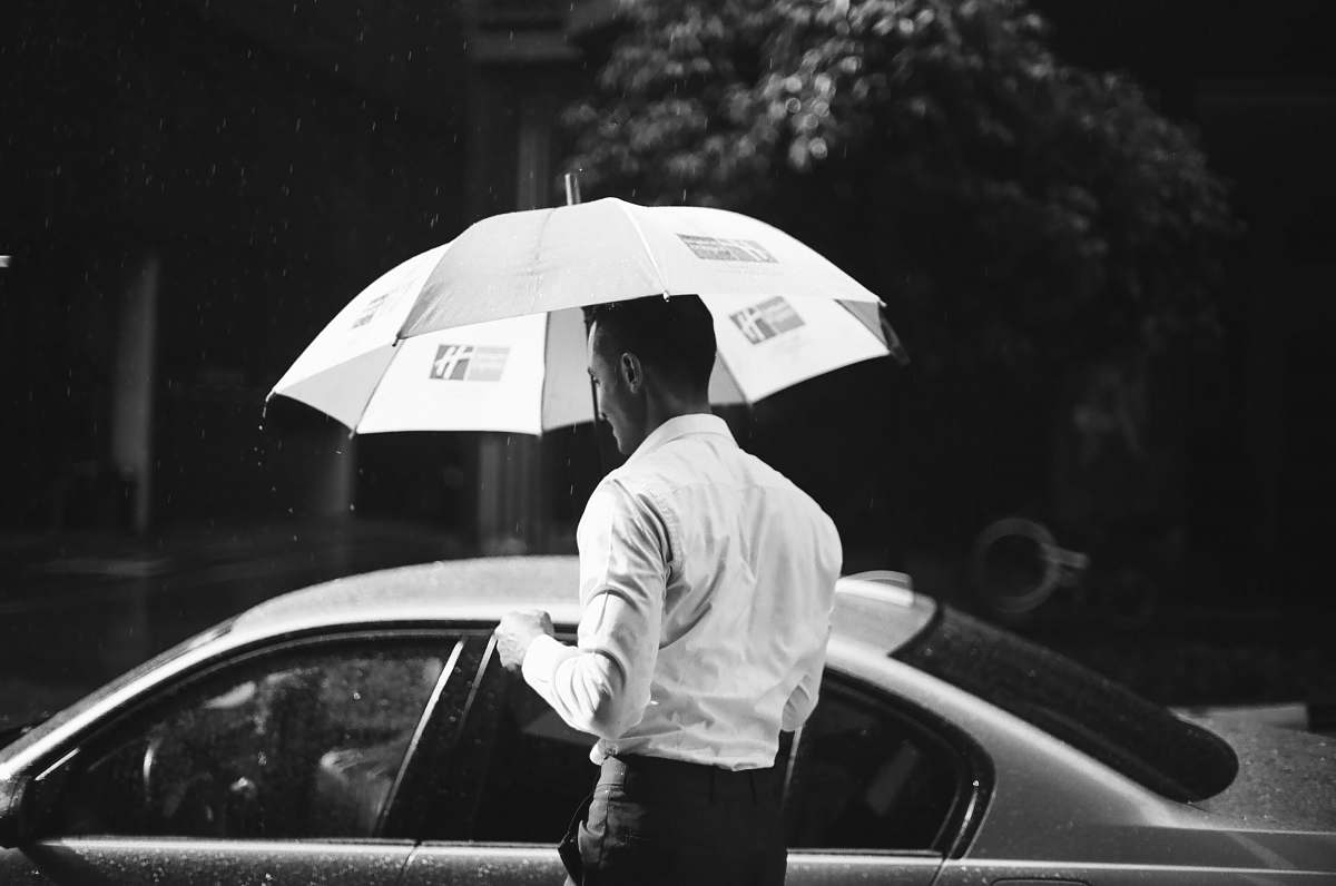 human grayscale photography of man holding umbrella in front of sedan black-and-white