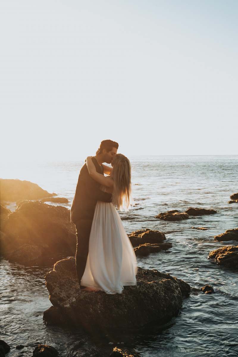 couple man and woman kissing on top of gray rock at beach kissing