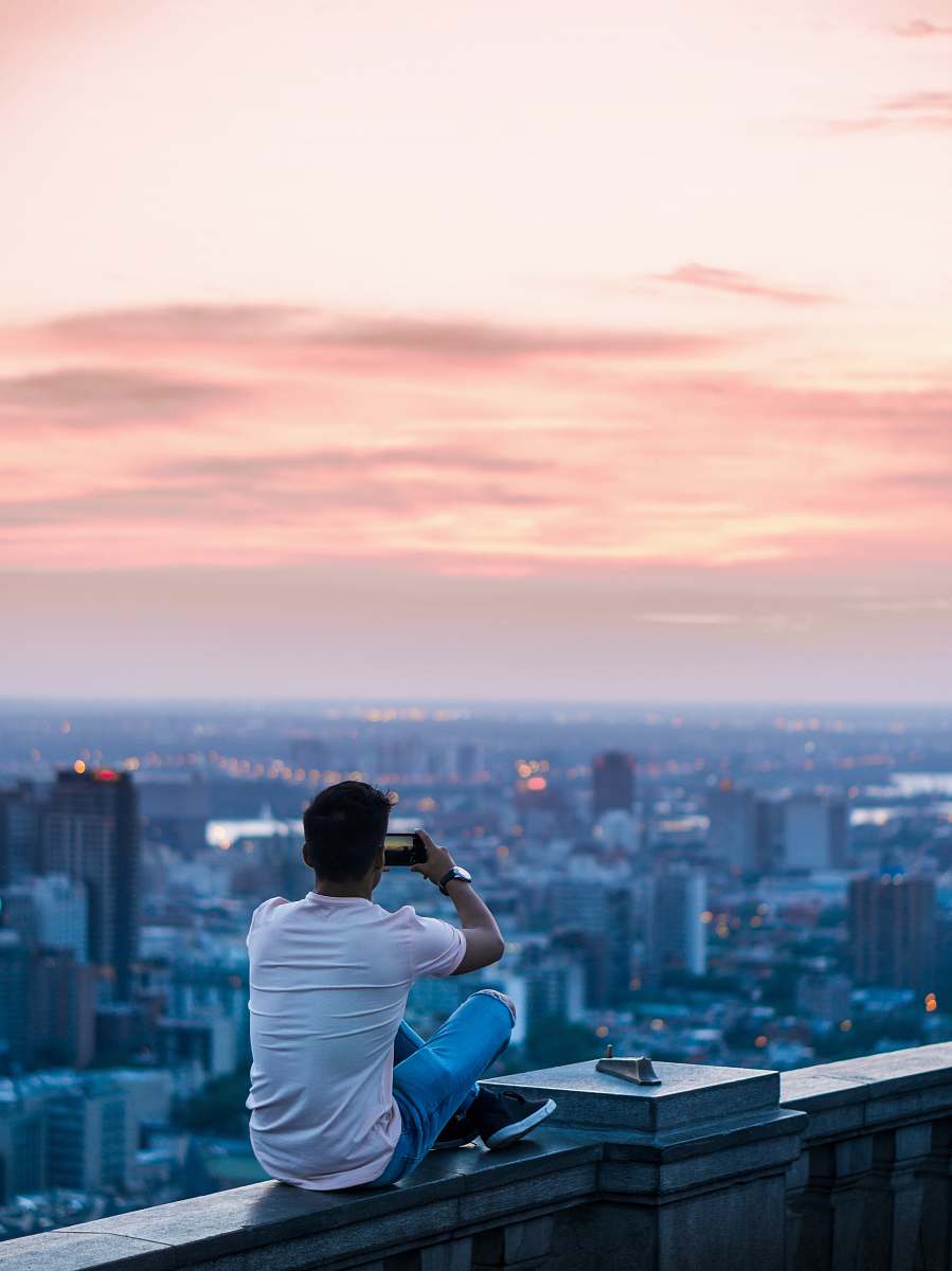 person person sitting on edge of building taking photo of city during daytime human