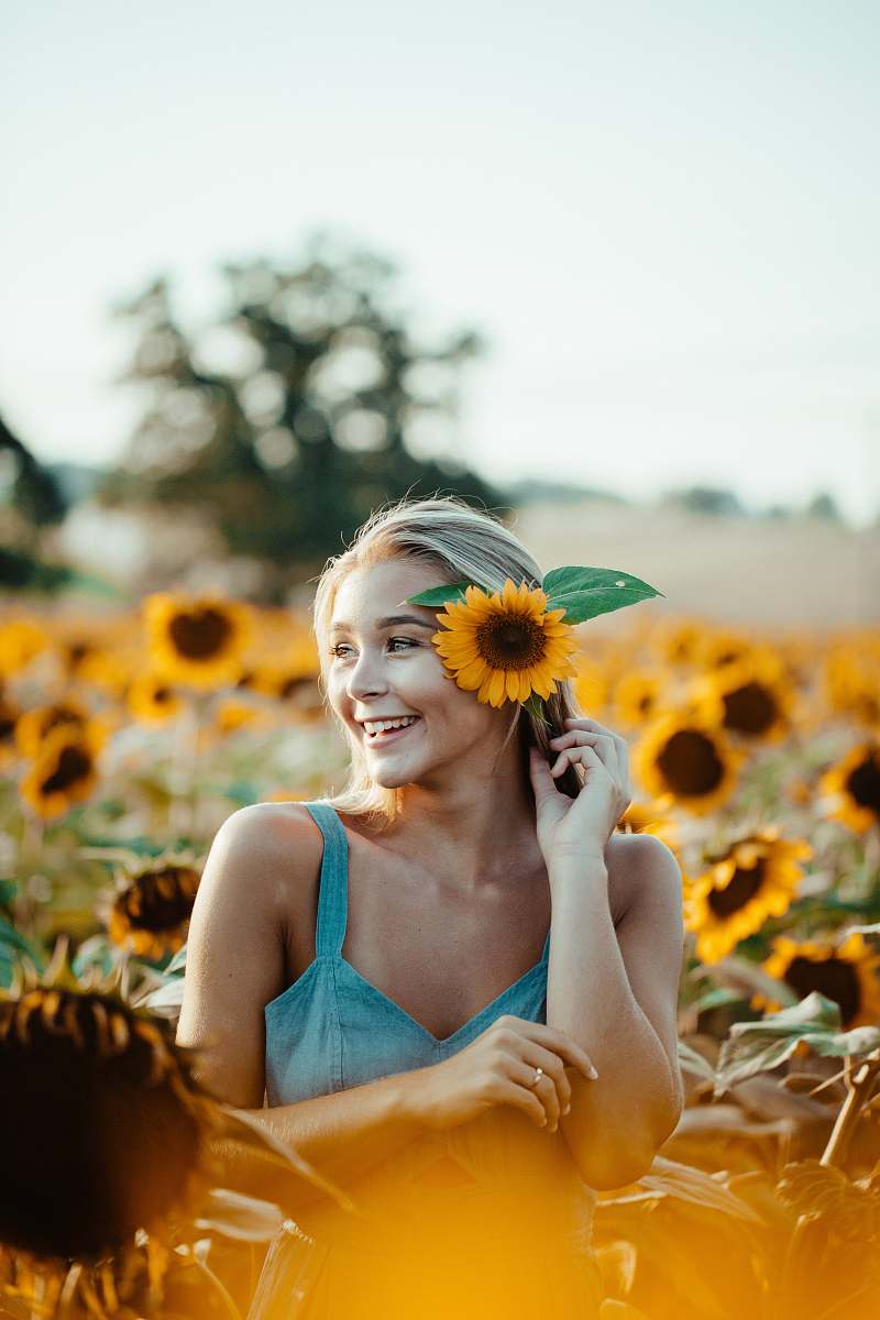 human woman in blue sleeveless dress holding sunflower placing in ear person