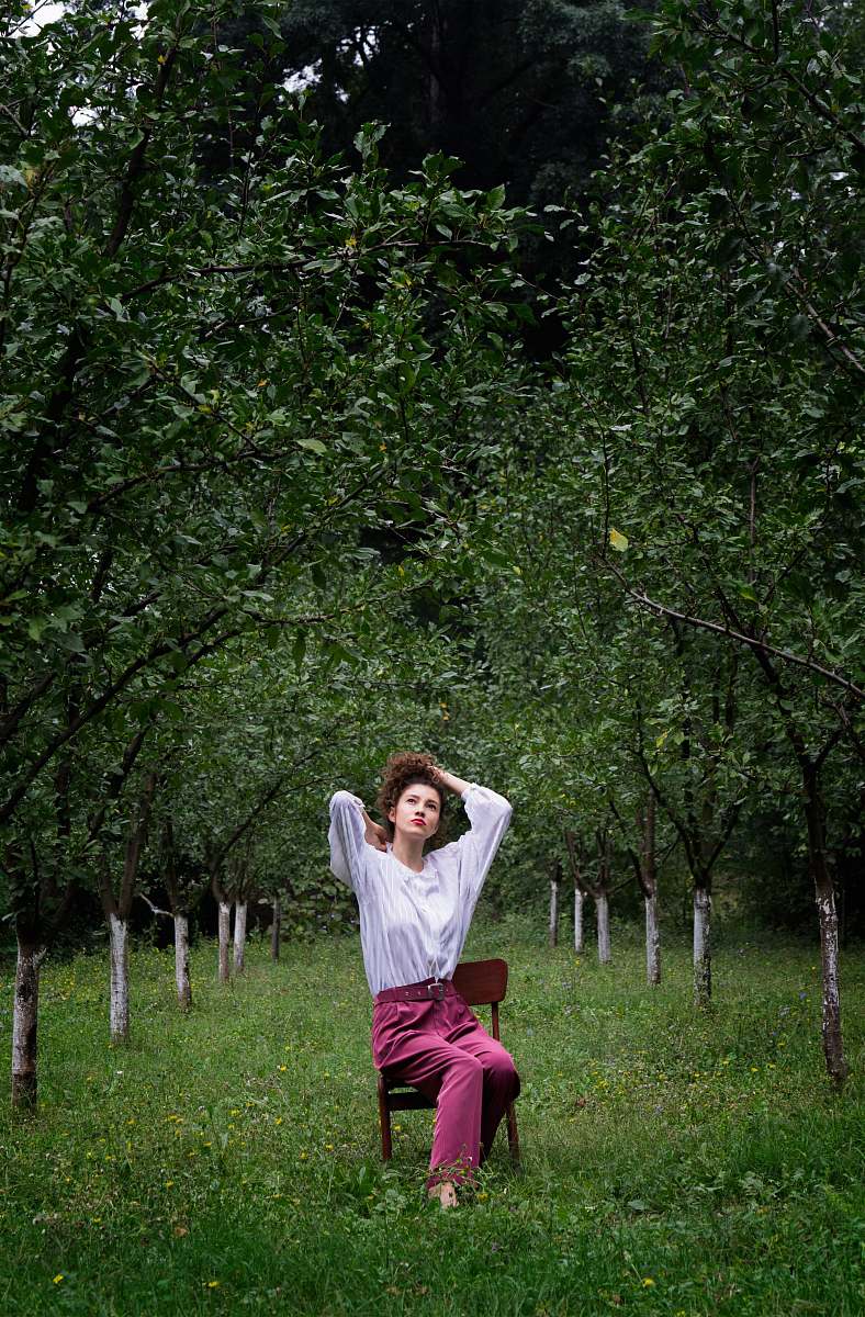 human woman sitting on brown armless chair while tying her hair under green trees at daytime person