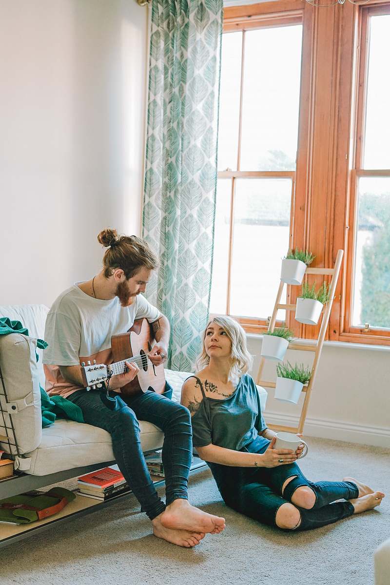 person man sitting on sofa playing guitar looking at girl sitting on the ground near window inside room people