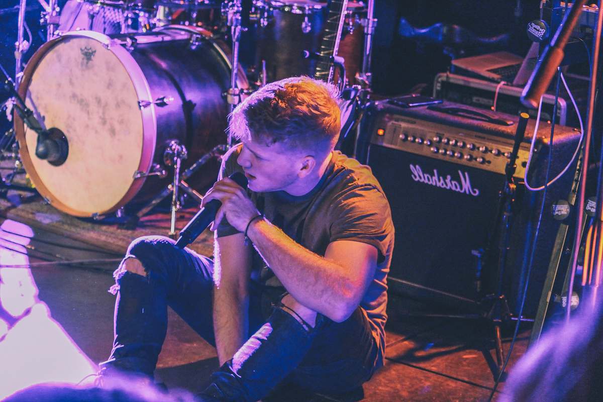 human vocalist man holding microphone sitting in front of Marshall guitar amplifier near drum kit person