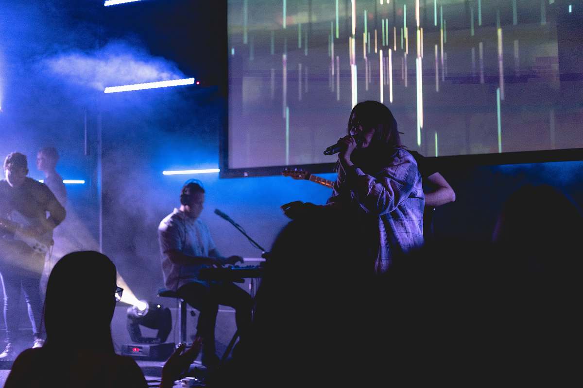 human boy singing with microphone beside man playing guitar, near man playing electronic keyboard, and man playing bass guitar on stage musician