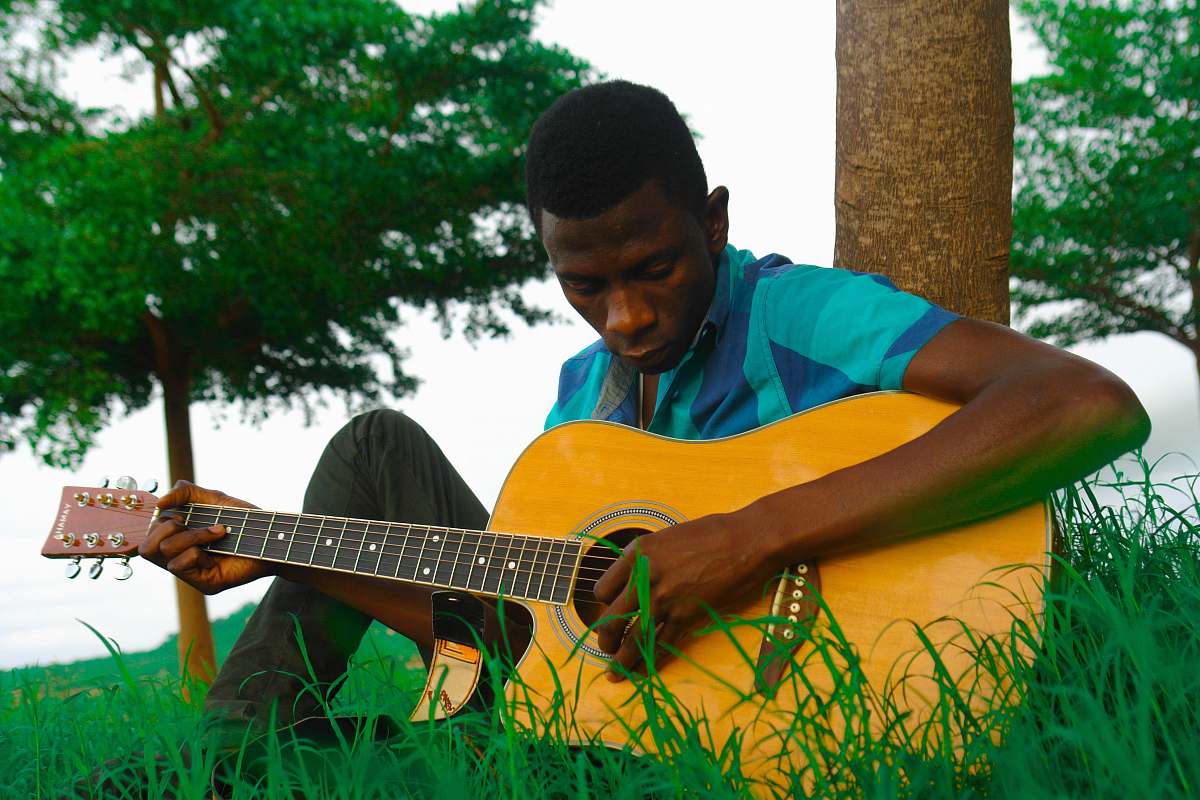 human man playing natural finish single-cutaway acoustic guitar while sitting on green grass near brown tree trunk during daytime people
