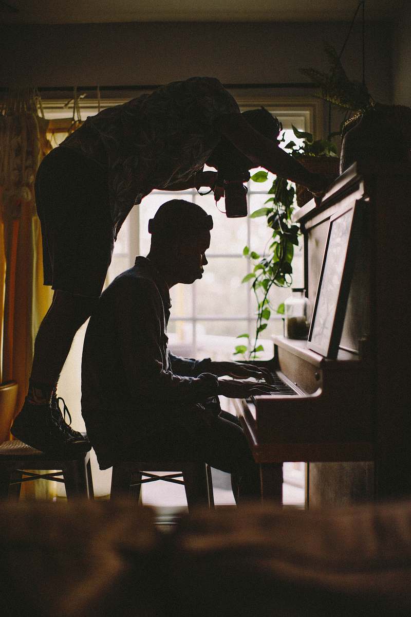 Immerse yourself in a captivating scene of creativity and artistry, featuring a serene individual passionately playing the piano. A photographer stands on a stool, capturing the delicate movement of the pianist's hands as they glide across the keys. This evocative image, set within a softly illuminated room, evokes a sense of inspiration and collaboration that is perfect for use in music-themed projects, artistic publications, or promotional materials for creative workshops.